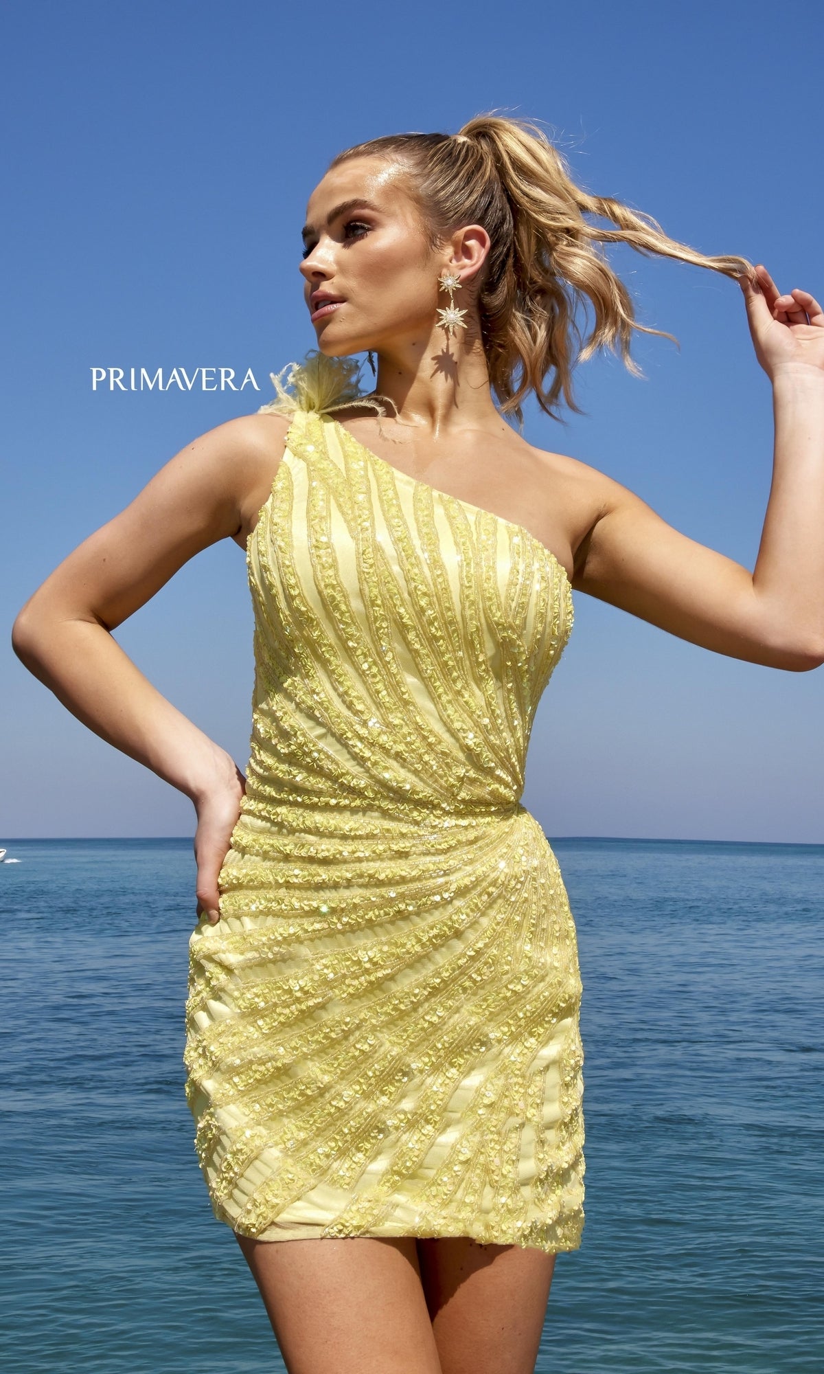 A woman stands by the ocean under a clear blue sky, wearing the One-Shoulder Feathered Beaded Cocktail Dress 4004.